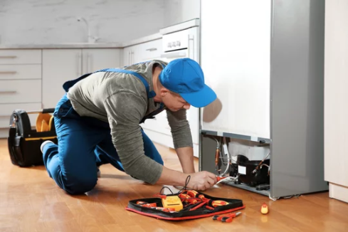 Technician performing maintenance on a refrigerator unit in a bright, professional service environment, demonstrating hands-on repair and diagnostic work with tools and equipment.